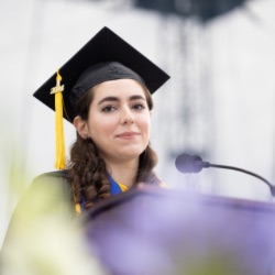 A young woman with long, dark hair wears a black graduation cap with a yellow tassel and gown. She stands at a podium with a microphone, appearing to give a speech. The background is blurred, emphasizing her as the focal point.